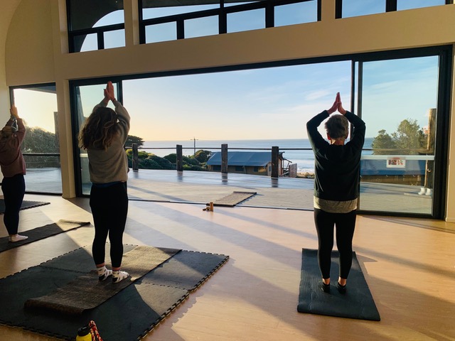 Two guests in a mountain pose on yoga mats on a timber floor undercover through overlooking through floor to ceiling window across the shoreline with a view - guests now have a new way to enjoy the Great Ocean Road at the BIG4 Pisces Apollo Bay caravan park