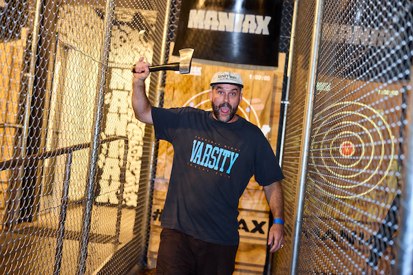 A man holding up an Axe ready to throw an axe in a indoor sporting lane divided with cyclone fencing each side at a Maniax venue. He's happy because he scored a free hour of axe throwing using the free code Maniax