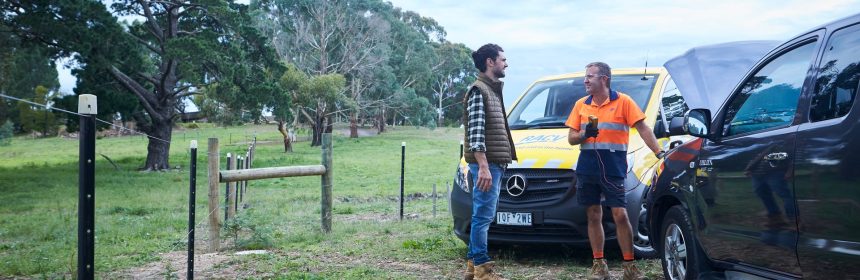 A man at the side of the road by his red fourbie looks happy to see an RACV technician standing in front of a yellow RACV mechanic van