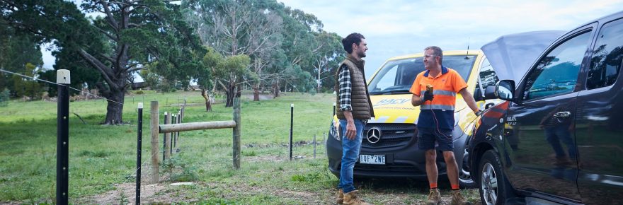 A man at the side of the road by his red fourbie looks happy to see an RACV technician standing in front of a yellow RACV mechanic van