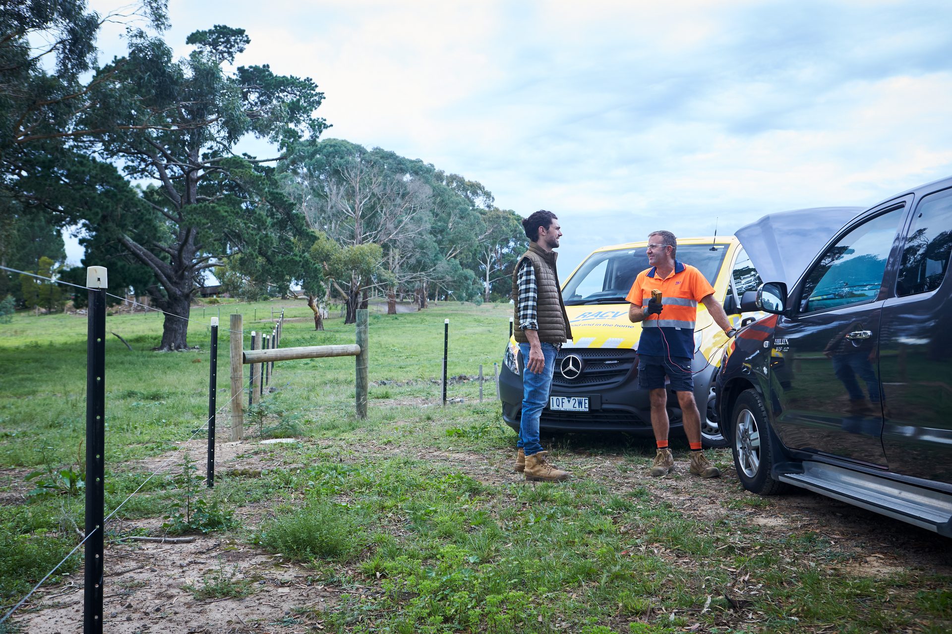 A man at the side of the road by his red fourbie looks happy to see an RACV technician standing in front of a yellow RACV mechanic van
